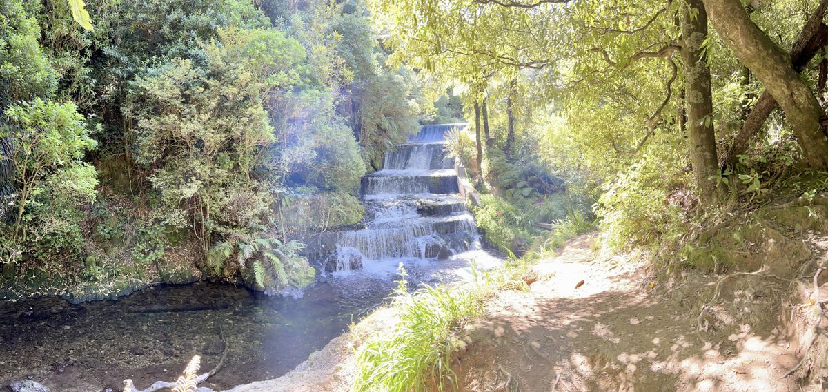 A cascading waterfall flows over multiple tiers, surrounded by lush, green foliage. Sunlight filters through the trees, creating a dappled effect on the ground.