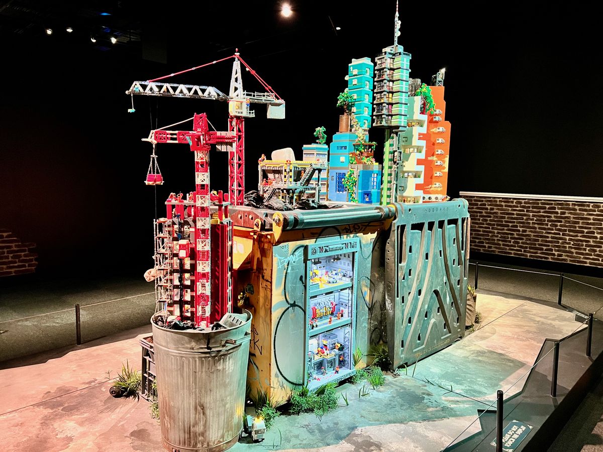 Model skyscraper buildings and construction cranes built from colorful LEGO bricks, standing on an oversized trashcan and a rusted metal container, within a dimly lit exhibition space.