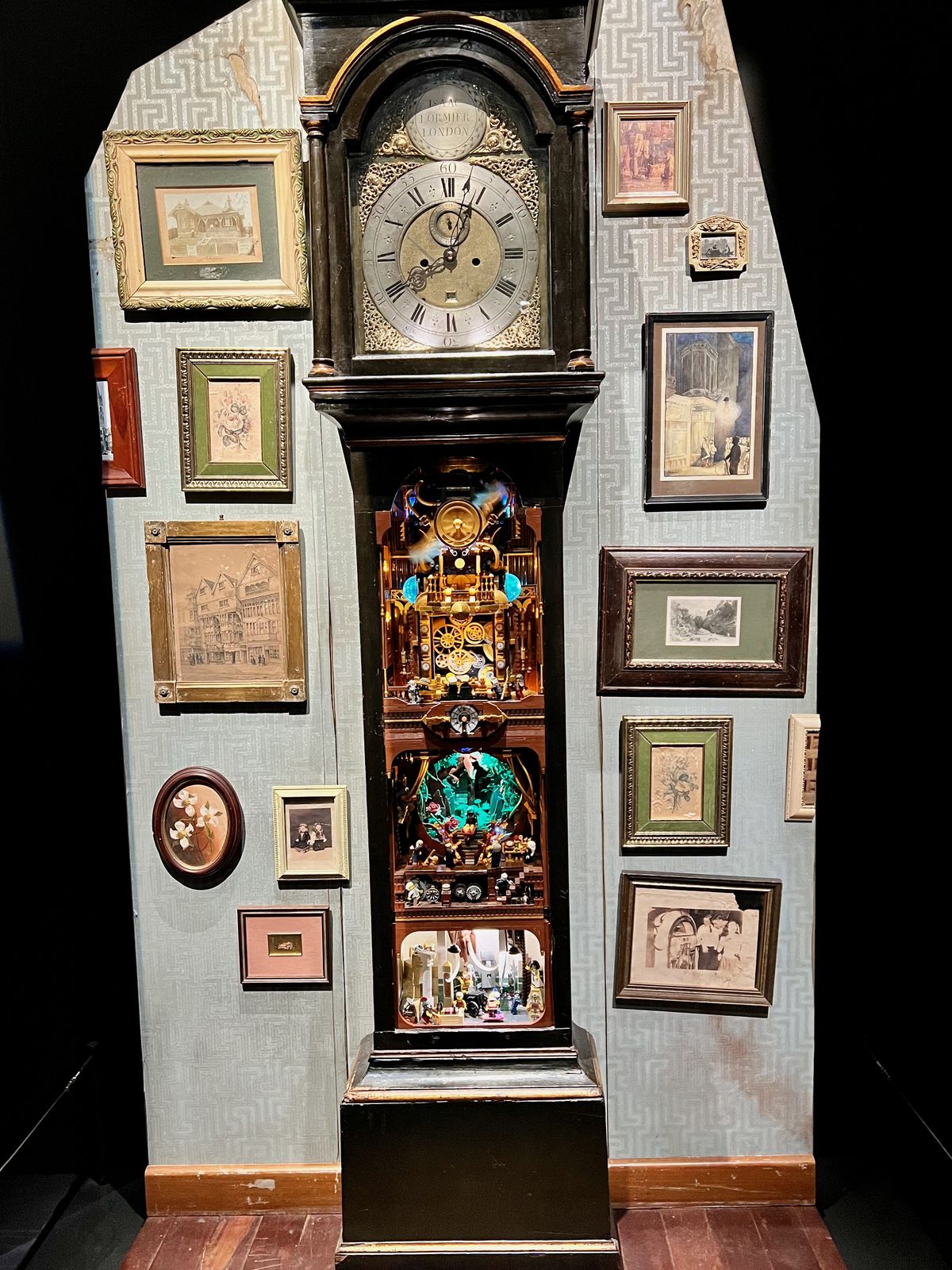 A scene built from colorful LEGO bricks within a rescued grandfather clock displaying intricate mechanical components, surrounded by framed artwork on a patterned wallpaper.