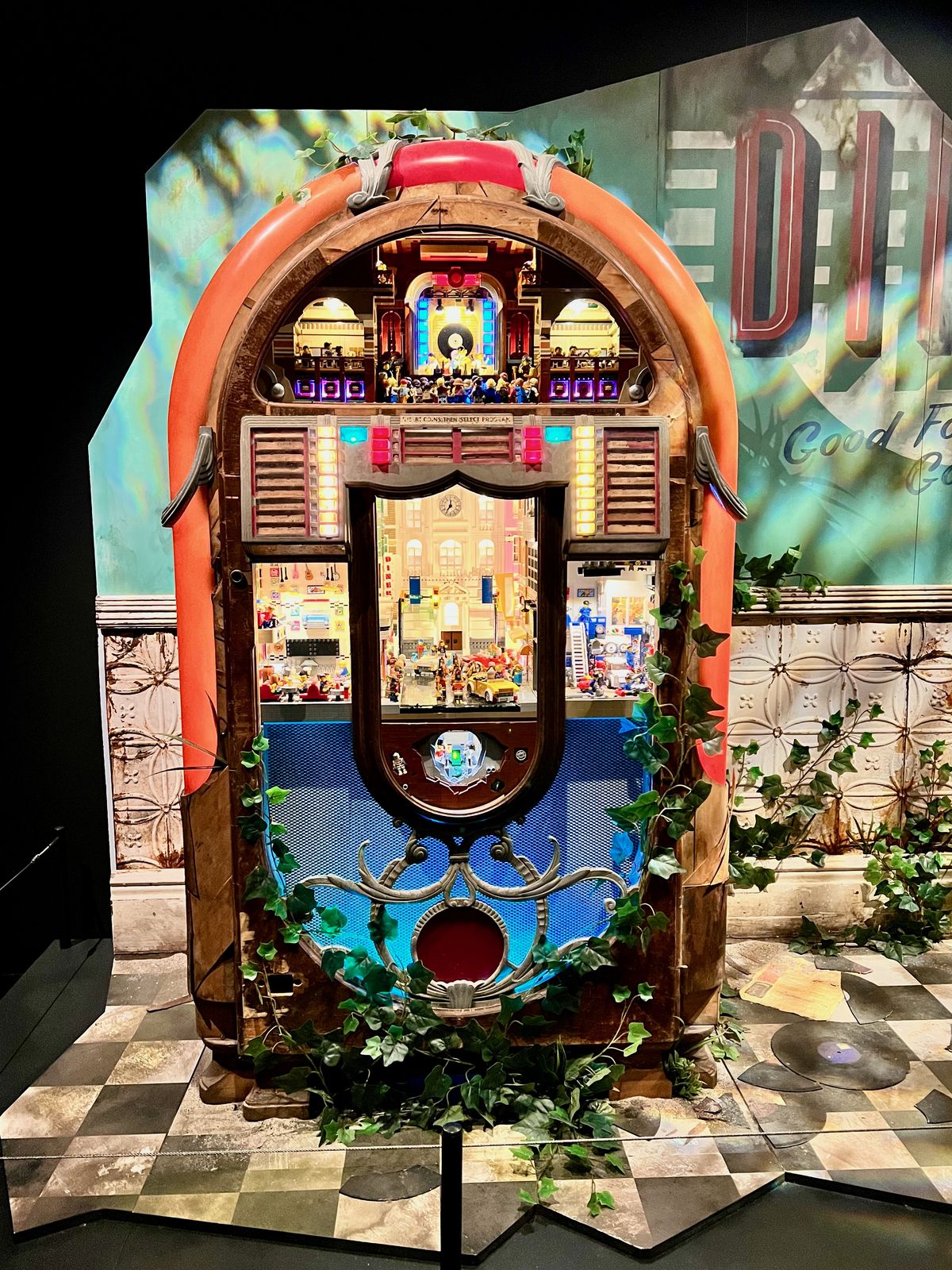 A colorful, retro jukebox stands in a decorated space, displaying small-scale figurines and intricate scenes inside, surrounded by a tiled floor and walls adorned with vintage posters and ivy.