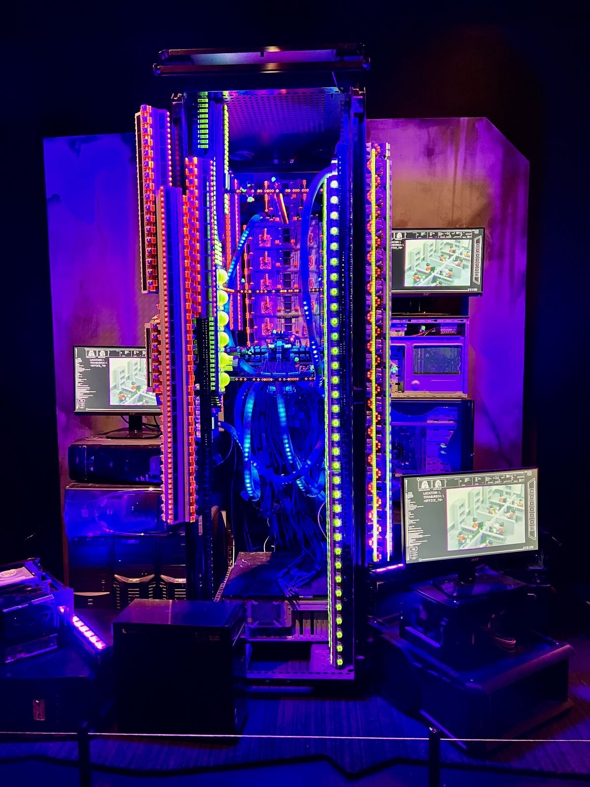 A server rack glows with vibrant LED lights, housing multiple electronic components and cables, surrounded by computer monitors displaying technical schematics, all set against a dark backdrop.