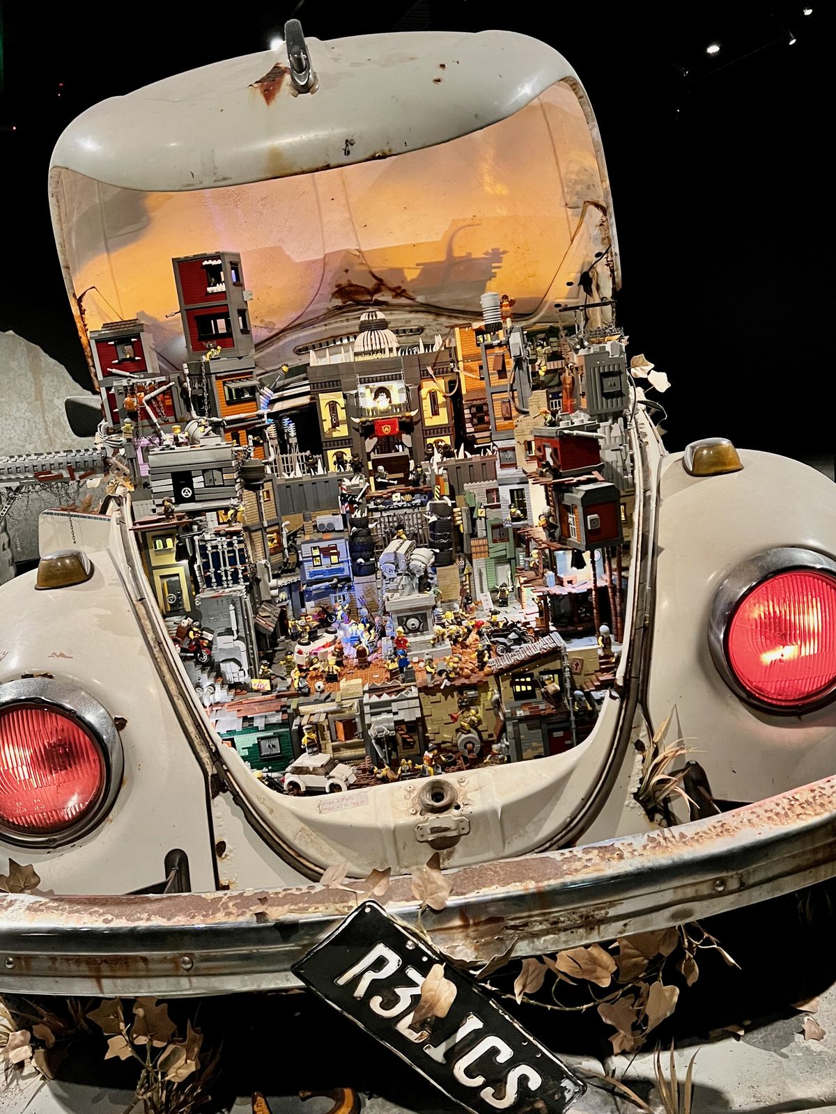 A miniature city made of toy blocks and figurines is crafted inside the open trunk of an old, rusty car with a tilted license plate reading 
