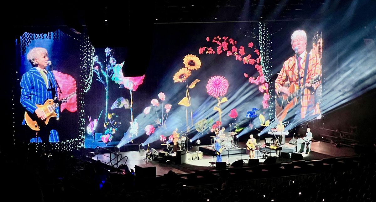 Split Enz perform on stage with guitars amid a vibrant floral-themed display, featuring large illuminated flowers. Spotlights and screen projections enhance the lively atmosphere in a concert setting.